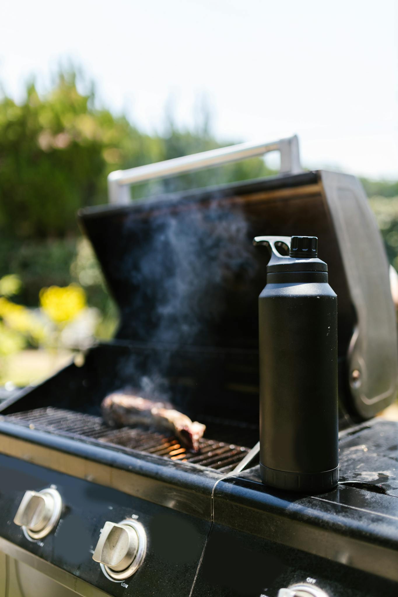 A close-up of a backyard barbecue grill with smoking meat and a tumbler, perfect for summer picnics.
